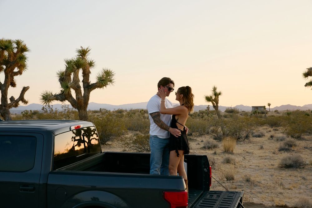 A couple embraces in the back of a pickup truck at sunset in the desert with Joshua trees in the background.