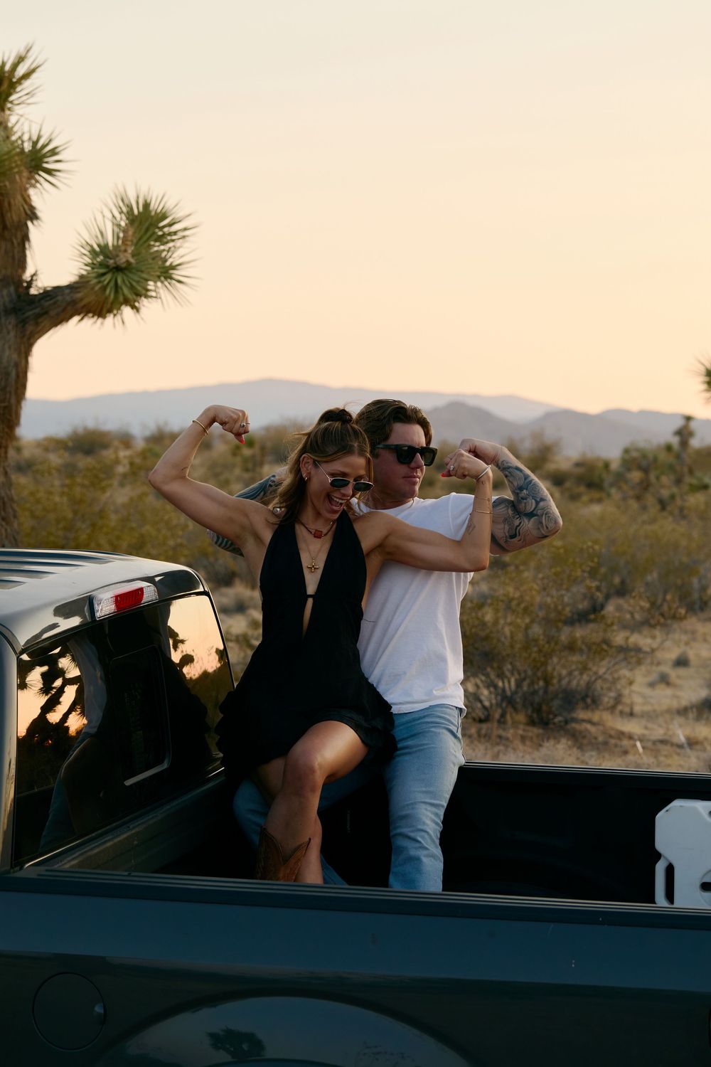A couple poses together in the back of a pickup truck in a desert landscape with mountains and a Joshua tree at sunset.