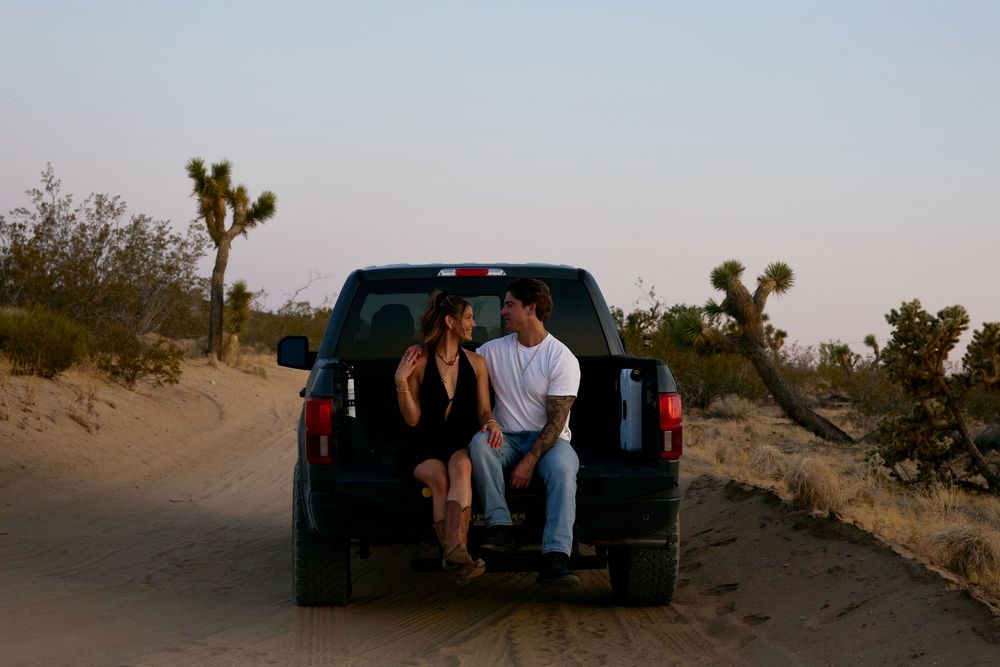 A couple sits in the back of a black pickup truck at sunset in a desert landscape with Joshua trees.