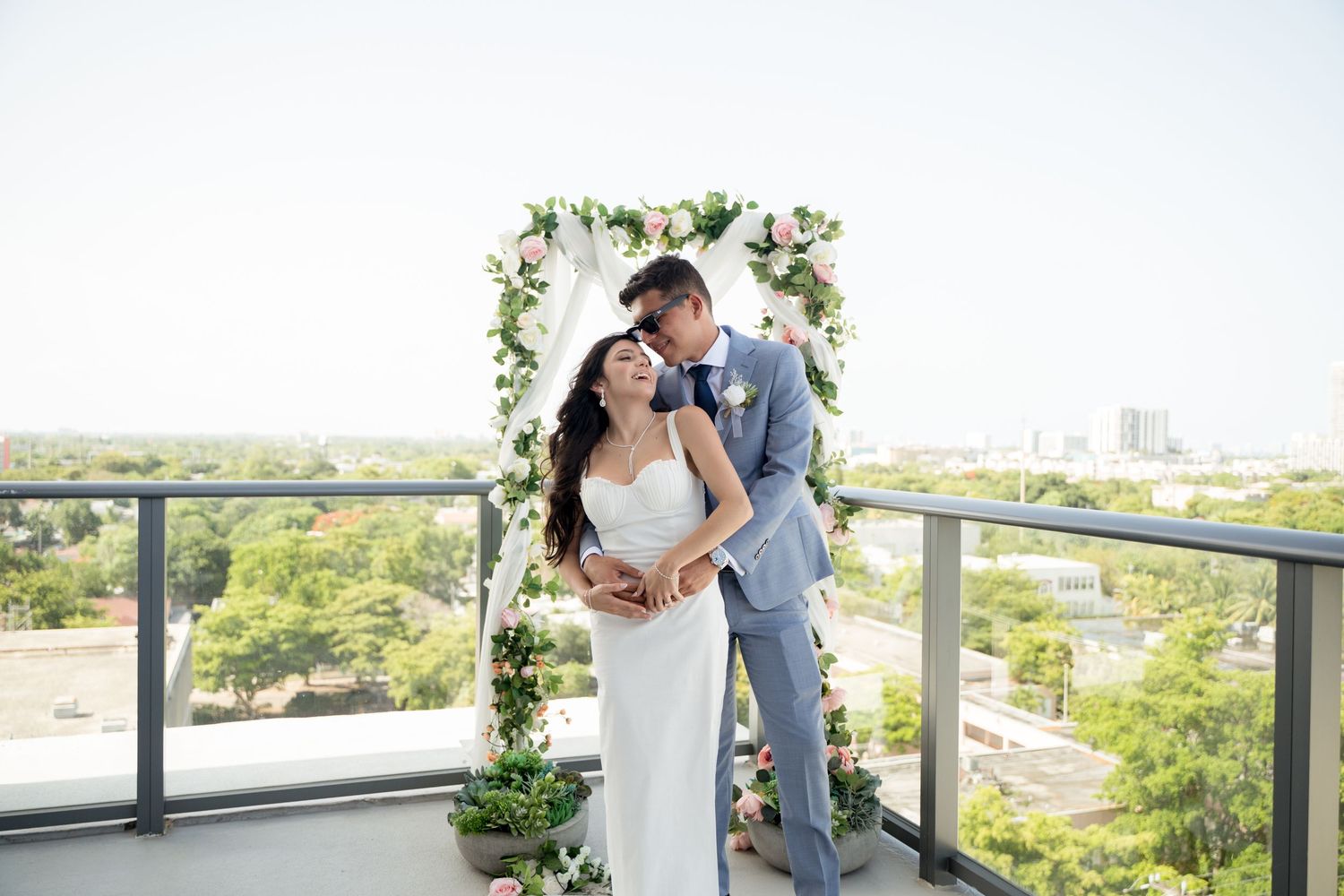 A couple shares an intimate moment on a balcony under a floral arch overlooking a scenic cityscape.