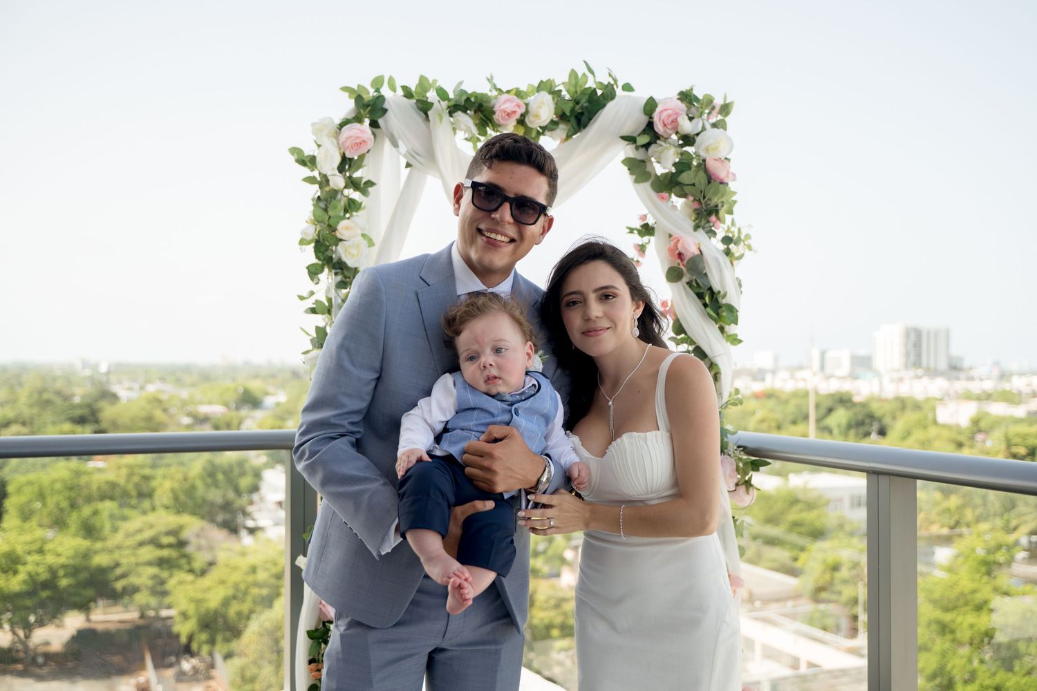 A couple and baby pose at an outdoor wedding ceremony with a floral arch and city skyline backdrop.