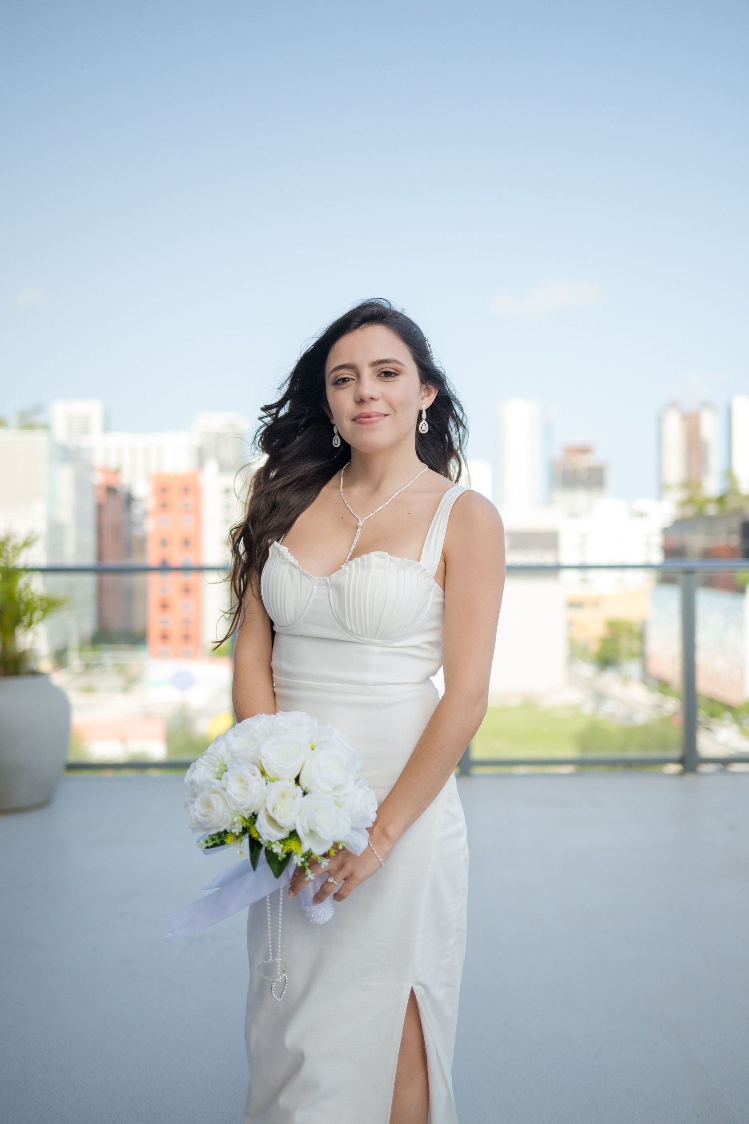 Bride in elegant white satin dress with spaghetti straps holds white floral bouquet on rooftop terrace with city backdrop.
