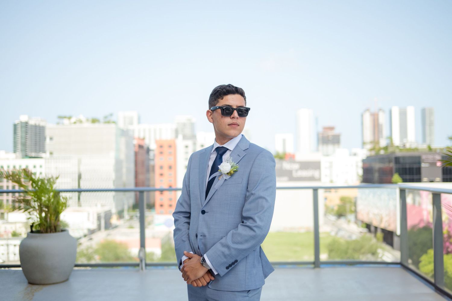 Figure in light blue suit and sunglasses stands on rooftop terrace with city skyline in background.