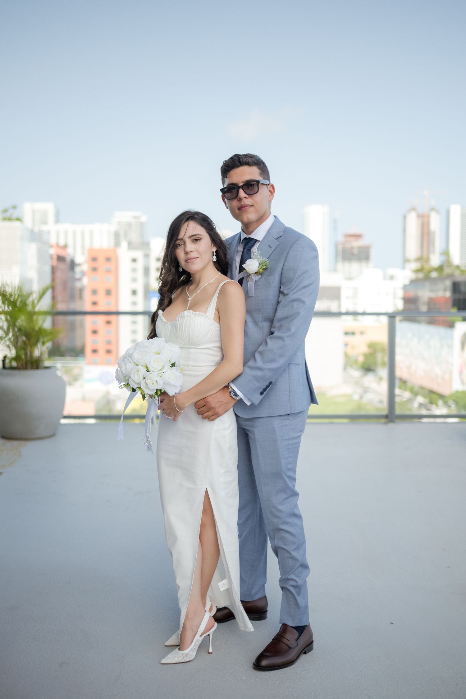 A bride in a white dress and groom in a gray suit pose together on a rooftop terrace with city buildings in the background.
