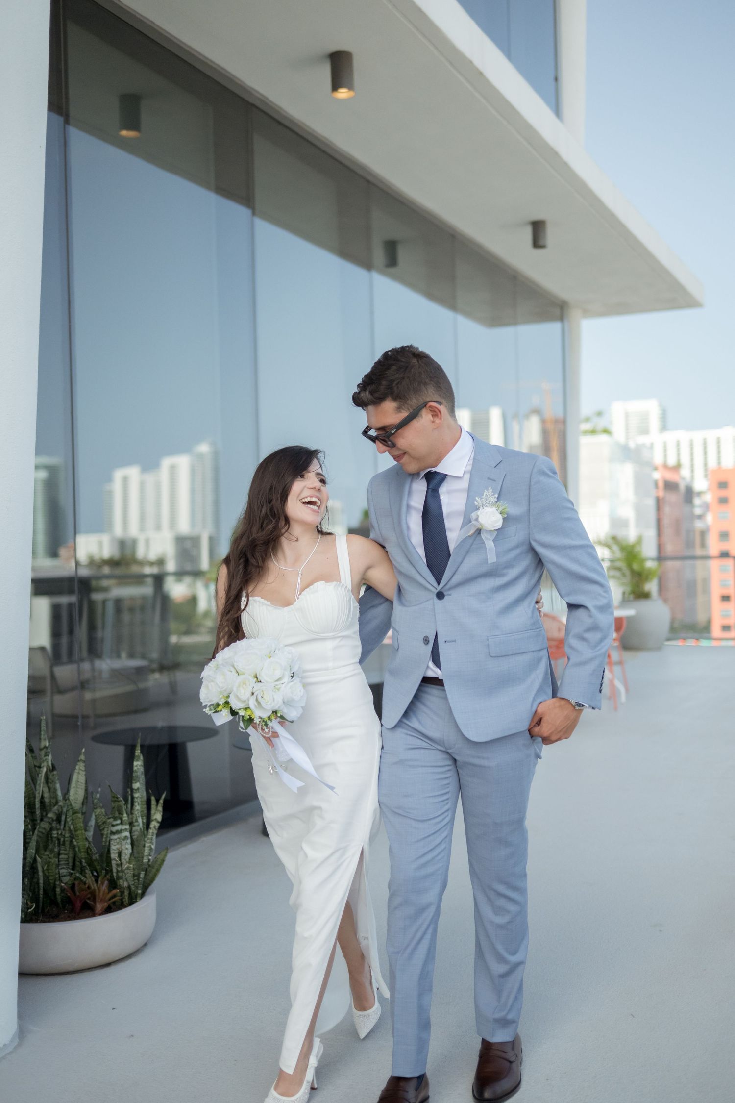 A couple in wedding attire walk together on a modern balcony with city skyline views in the background.