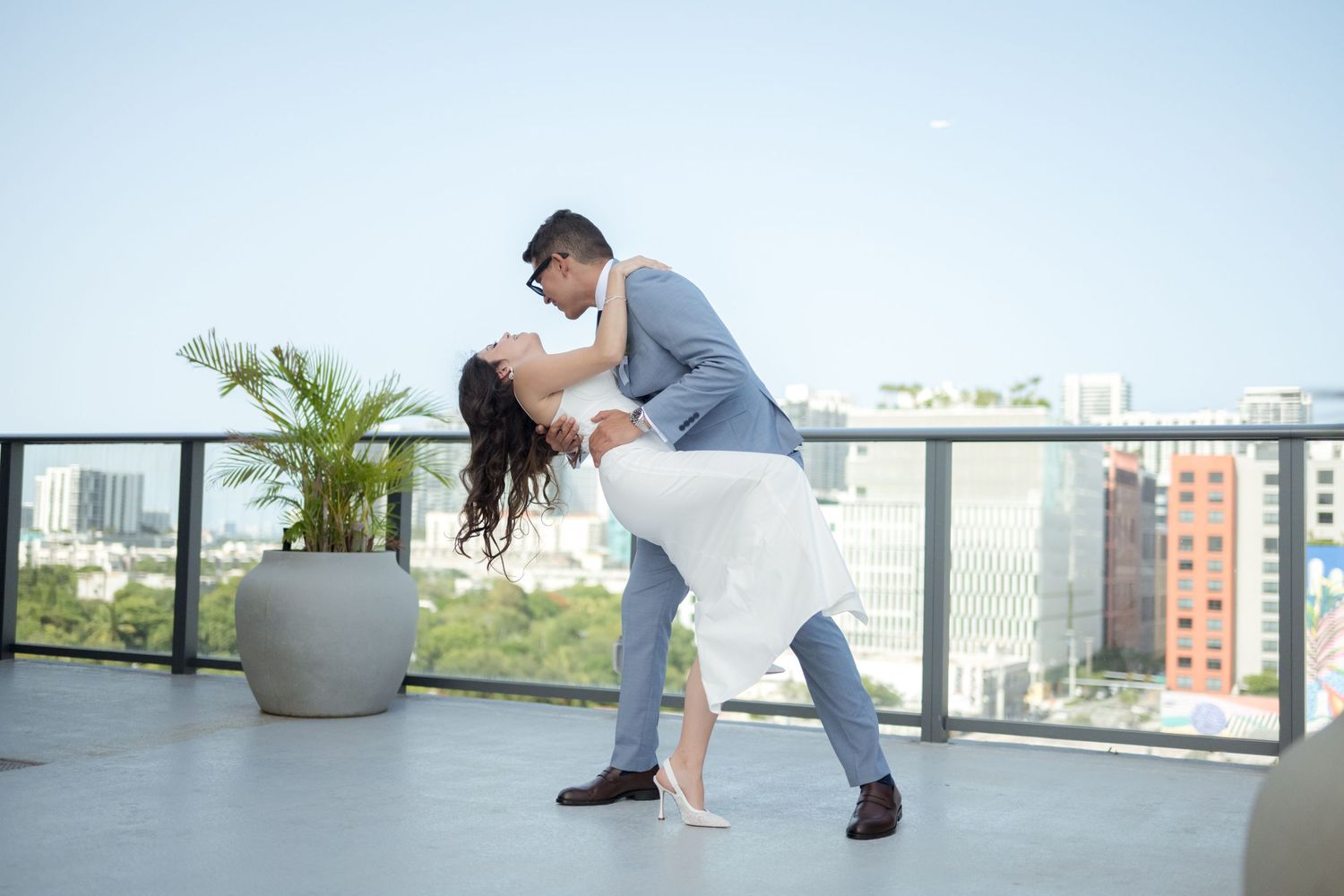 A couple dances together on a rooftop terrace overlooking city buildings with a potted palm plant in the background.