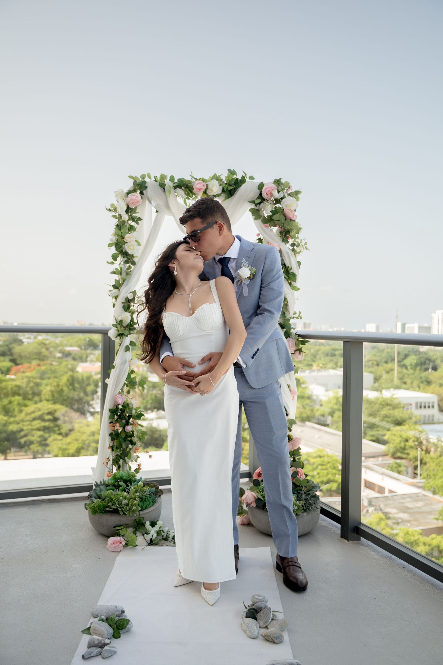 Newlyweds share a romantic kiss on a rooftop balcony decorated with a floral arch against an urban backdrop.