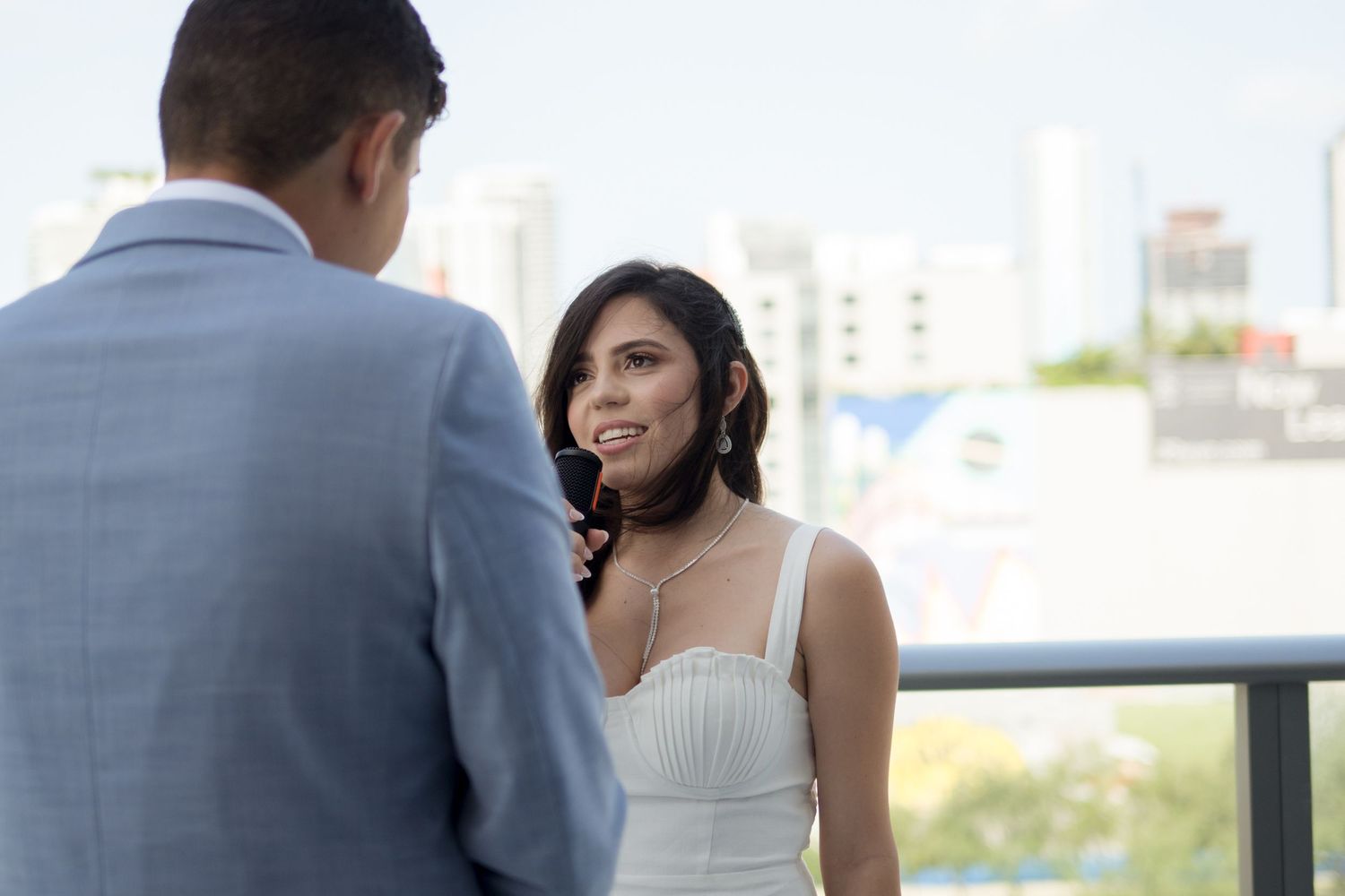 A couple in wedding attire shares an intimate moment on a balcony overlooking a city skyline.