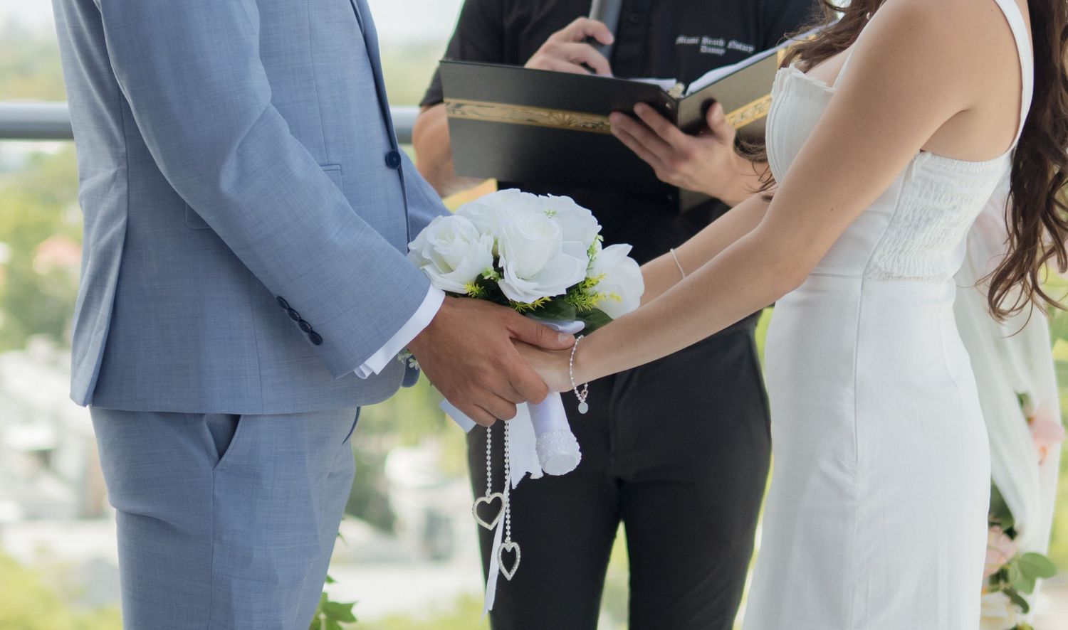 Wedding ceremony participants gather outdoors on a sunny day with white floral decorations and formal attire.