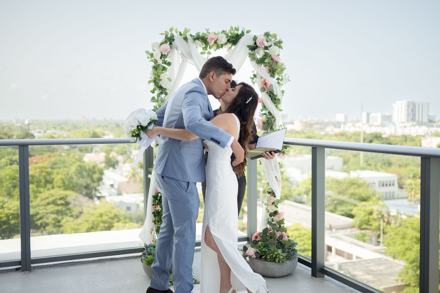 A romantic couple shares a moment under a floral wedding arch on a balcony overlooking a city landscape.
