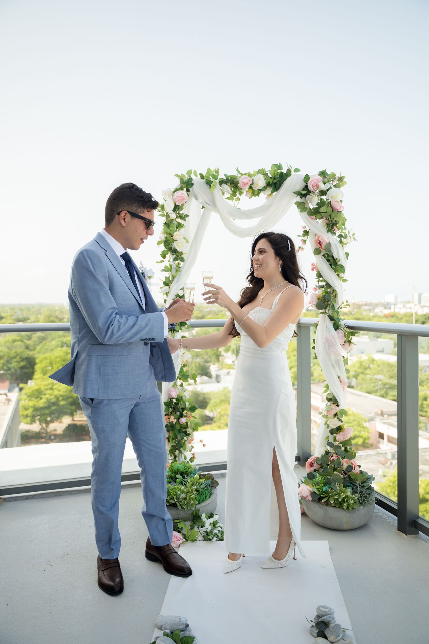A bride and groom exchange vows under a floral arch on an outdoor balcony overlooking a scenic view.