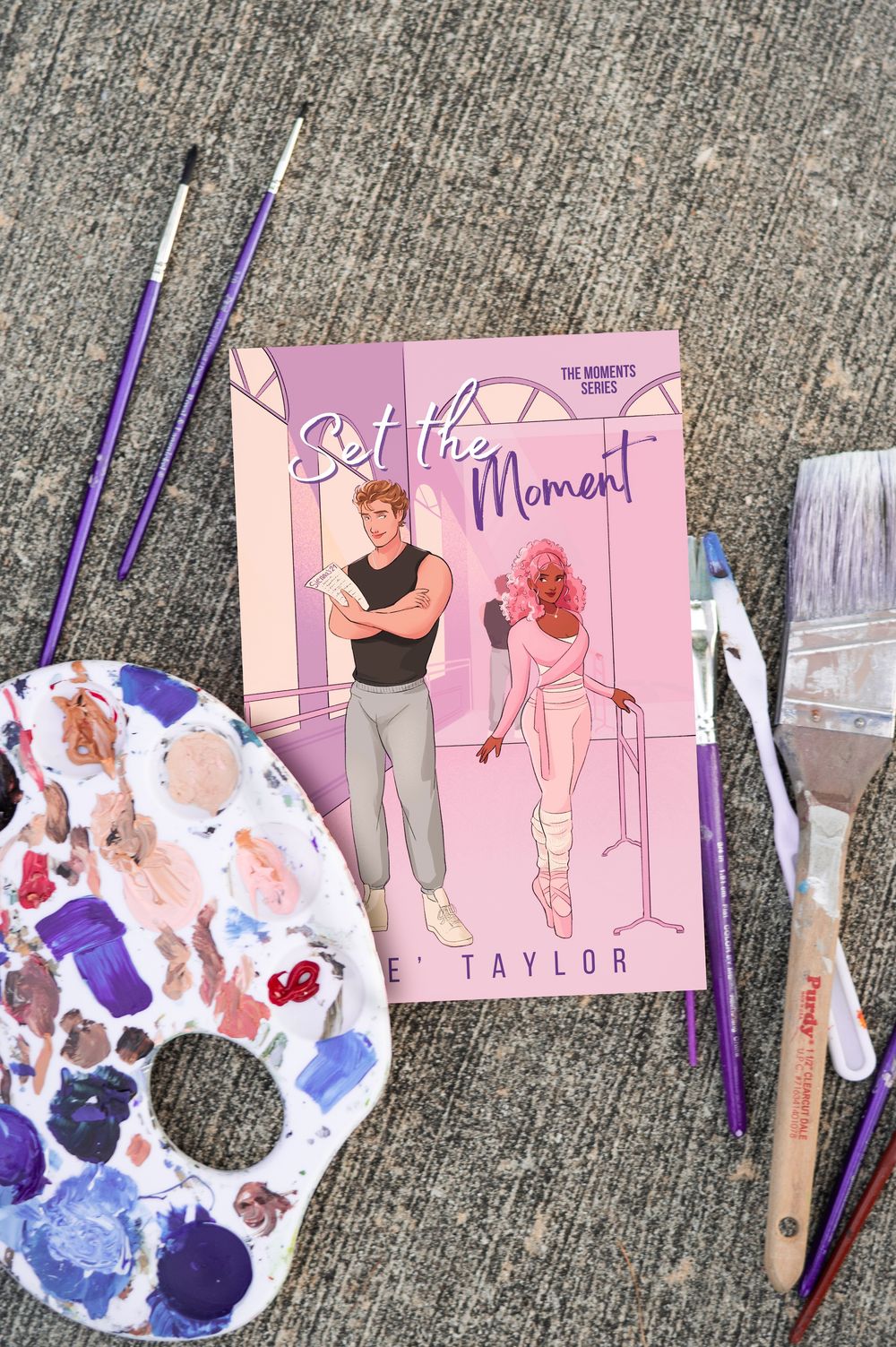 Art supplies and pink-themed illustrations are arranged on a concrete surface featuring watercolor paint palettes and brushes.