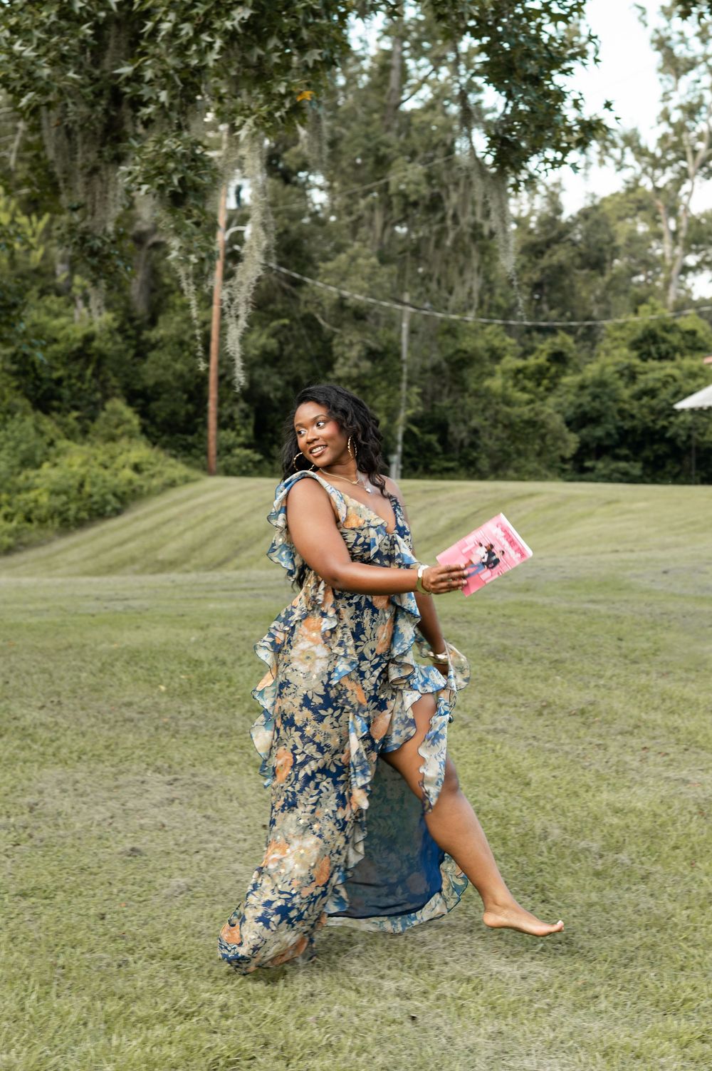 Someone in a floral dress poses outdoors on grassy lawn with trees in the background.
