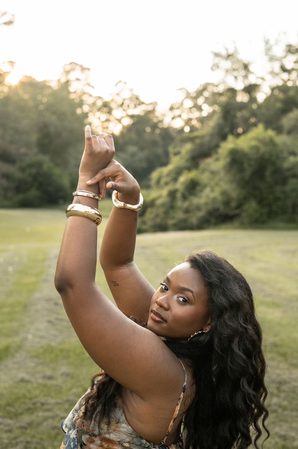 Person with long wavy dark hair and bracelets poses gracefully with raised arms in a sunny outdoor field at sunset.