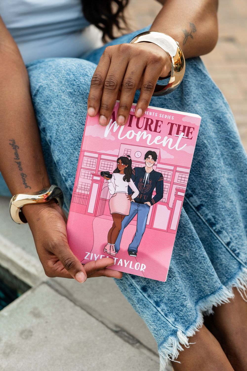 Person holding a pink novel with romantic book cover artwork while wearing distressed denim jeans.