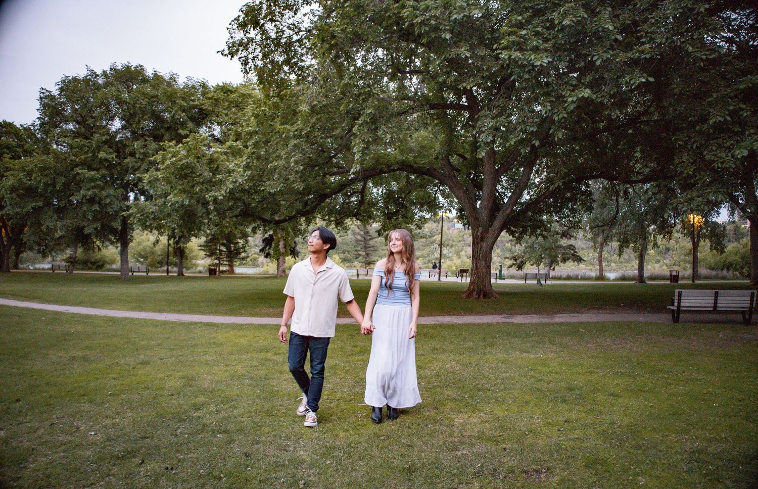 Five young women sitting on a bench outdoors, laughing together in casual jeans and tops with green trees behind them.