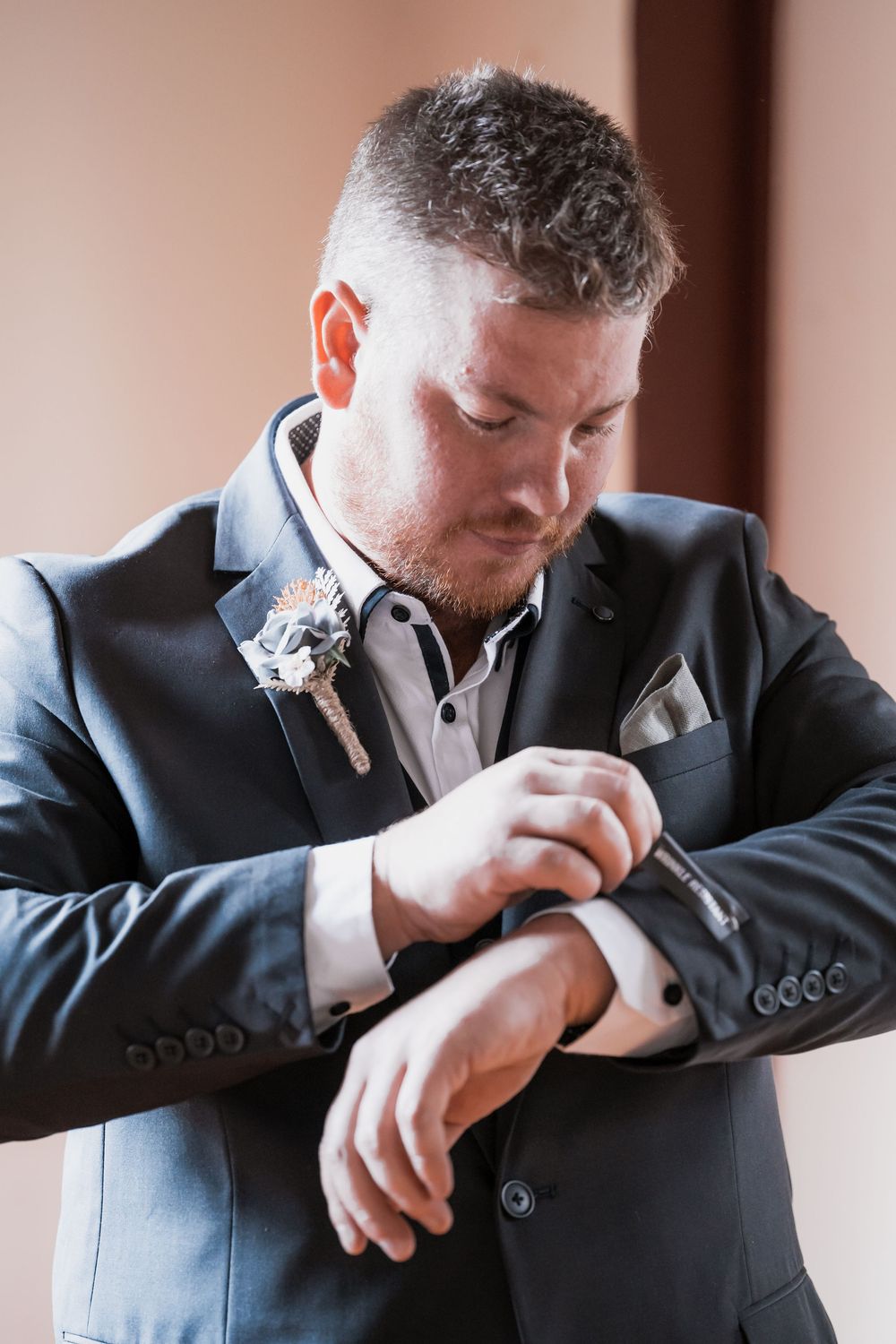 Groom in black suit adjusts his watch while getting ready on wedding day, with boutonniere pinned to lapel.