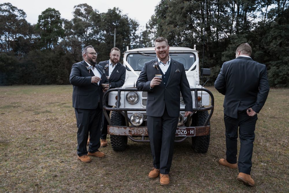 Professional groomsmen gather around vintage cars during outdoor wedding preparations on a grassy field.