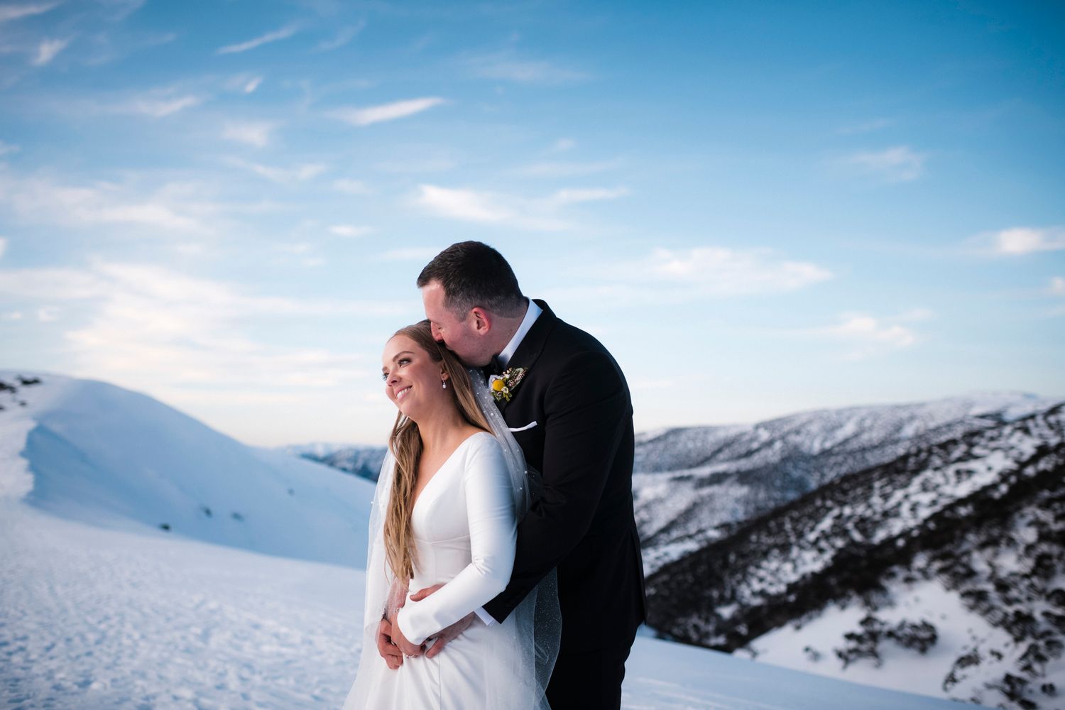 A couple in wedding attire embrace on a snowy mountaintop with snow-capped peaks and blue sky in the background.