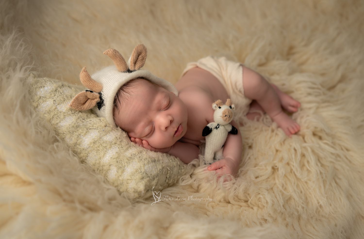 Sleeping newborn in cream outfit laying on fluffy white fur rug while holding small plush cow toy.