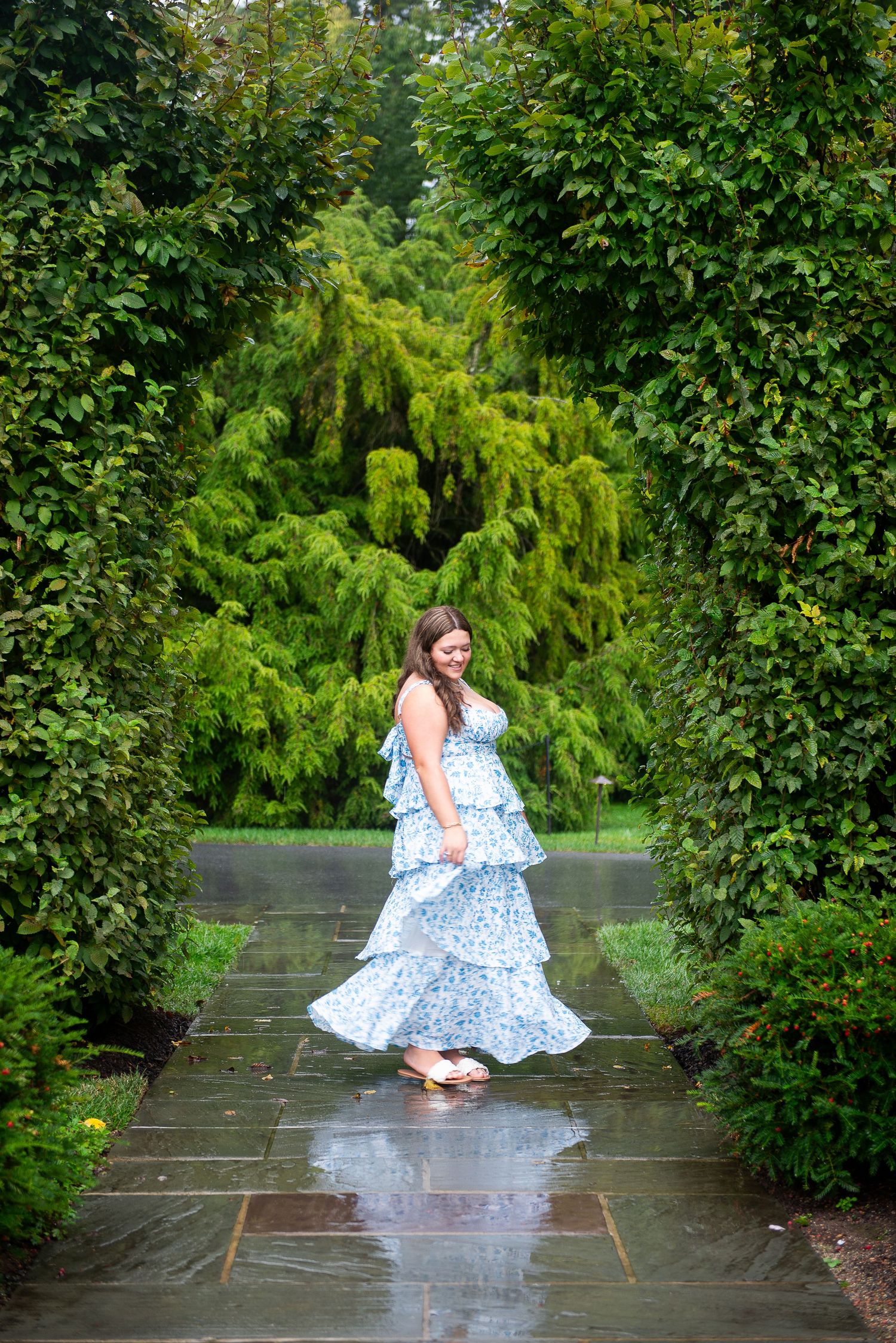 A figure in a blue floral dress crouches down on wet garden pavers surrounded by lush green foliage.