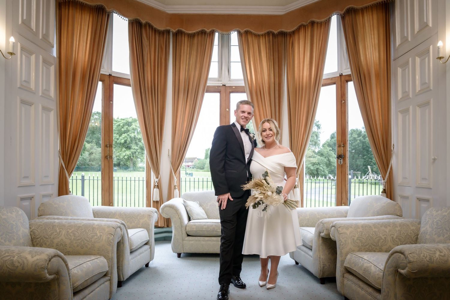 A couple stands together in an elegant room with tall windows, gold curtains and plush seating at a luxury venue.