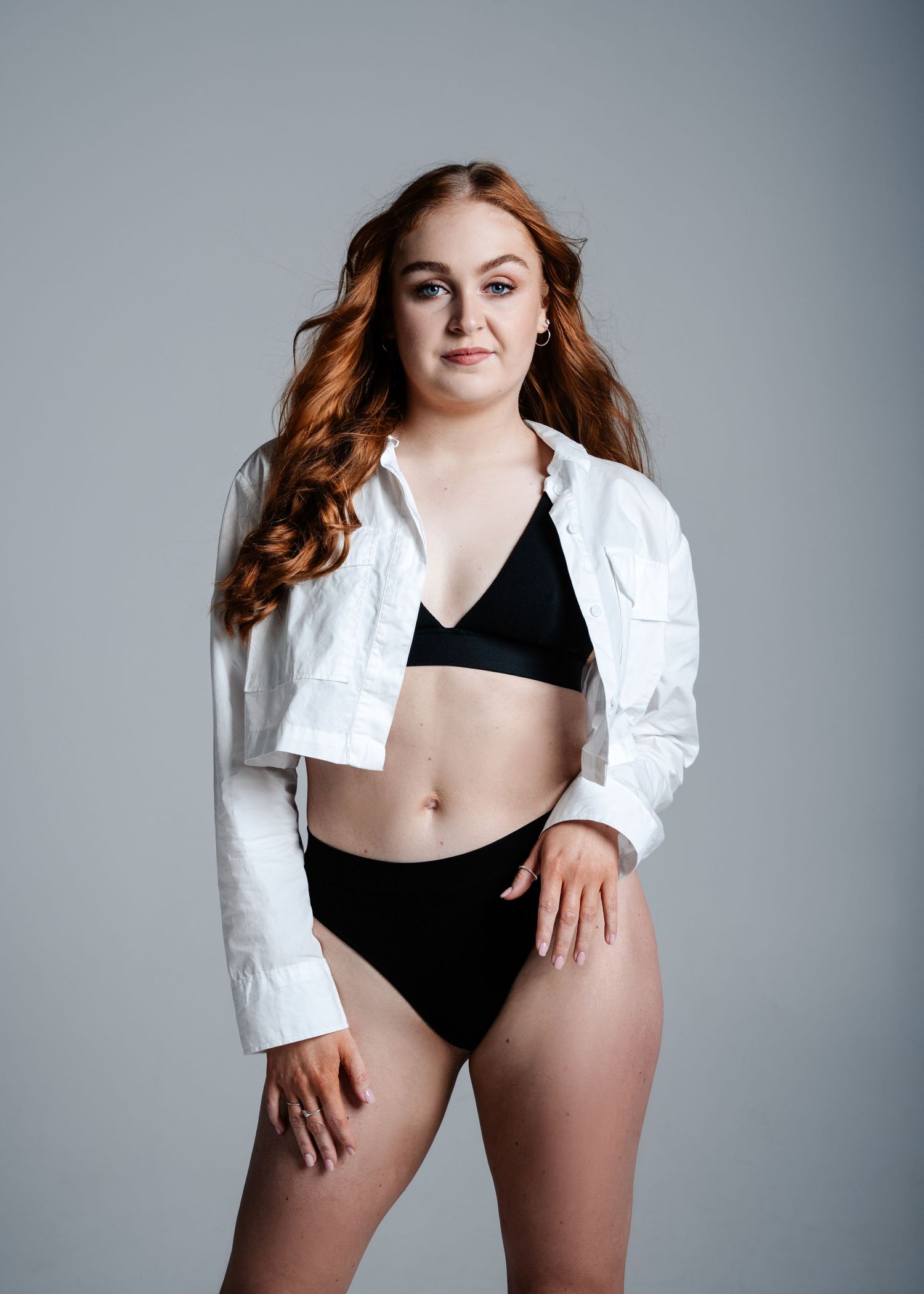 A model lounges in a chic black bathing suit and white blazer against a neutral grey background.