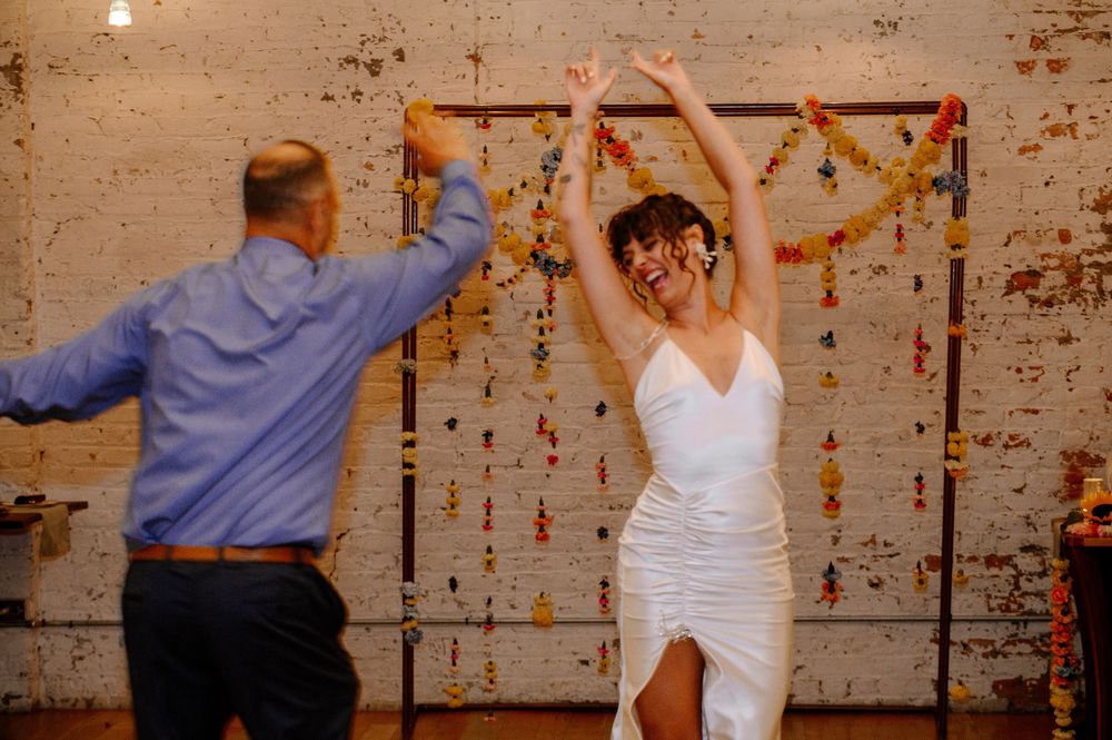 Wedding guests dance joyfully against a rustic brick wall backdrop decorated with dangling string lights.