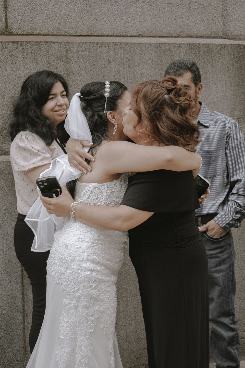 A bride in a white lace dress shares an emotional embrace at a wedding while others look on and smile.