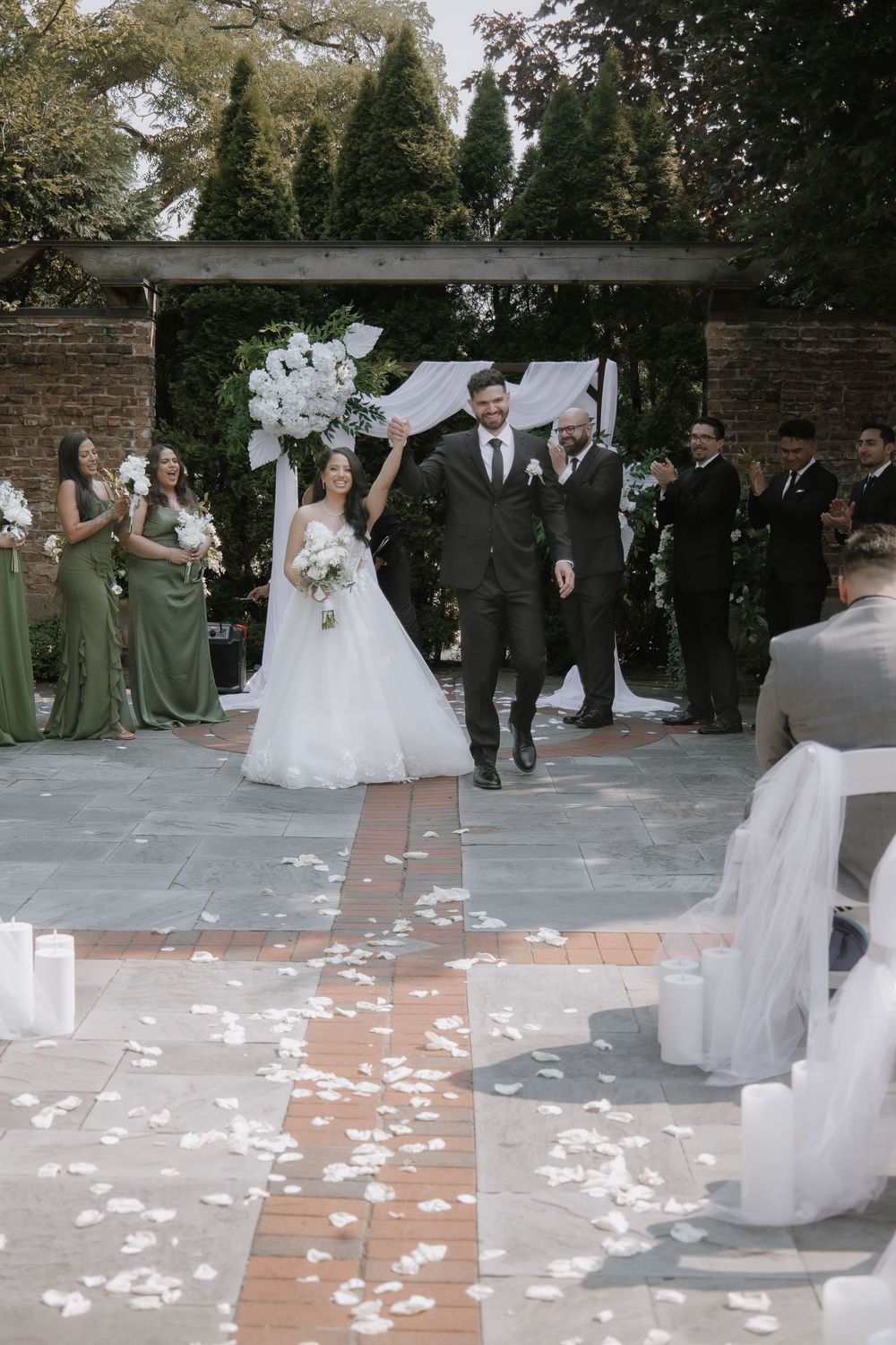 Elegant outdoor garden wedding ceremony with green-clad bridesmaids and floral arch beneath trees on brick pathway.