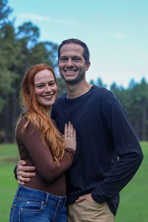 Happy couple embracing and smiling outdoors on a green lawn with pine trees behind them.