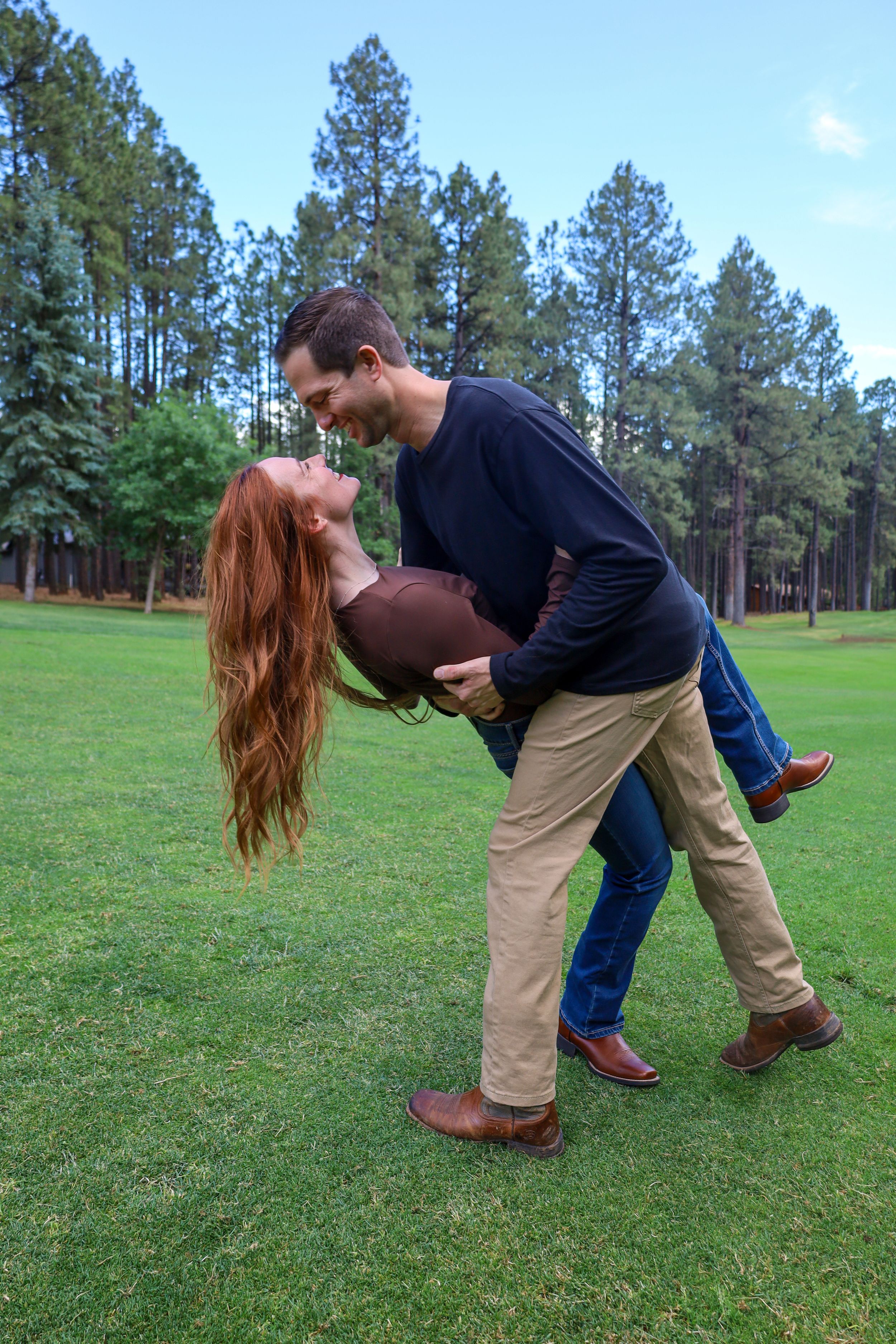 Happy couple laughing as man lifts woman on a lush green golf course with pine trees.