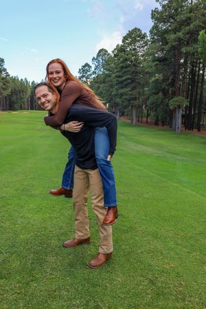 Smiling couple enjoying a playful piggyback ride on a green golf course with pine trees.