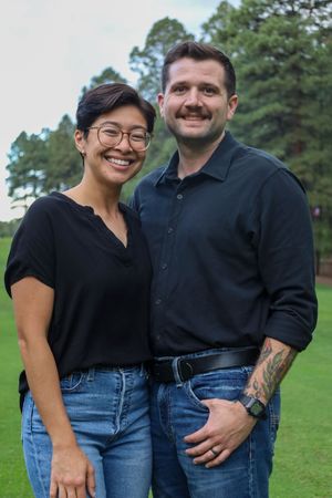 Smiling interracial couple posing together on a green lawn surrounded by tall pine trees.