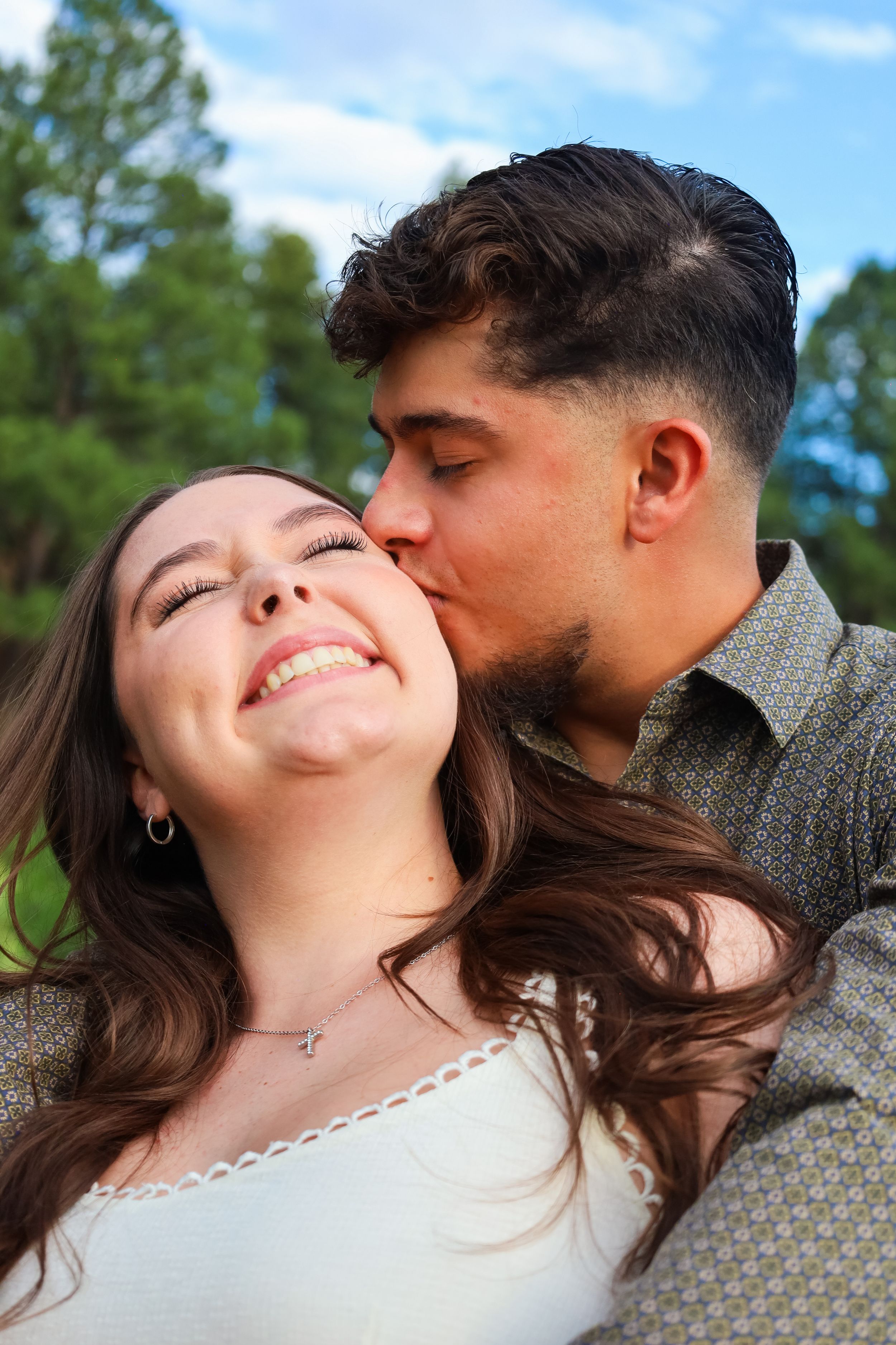 A young man kisses a smiling brunette woman on the cheek in an outdoor park setting.