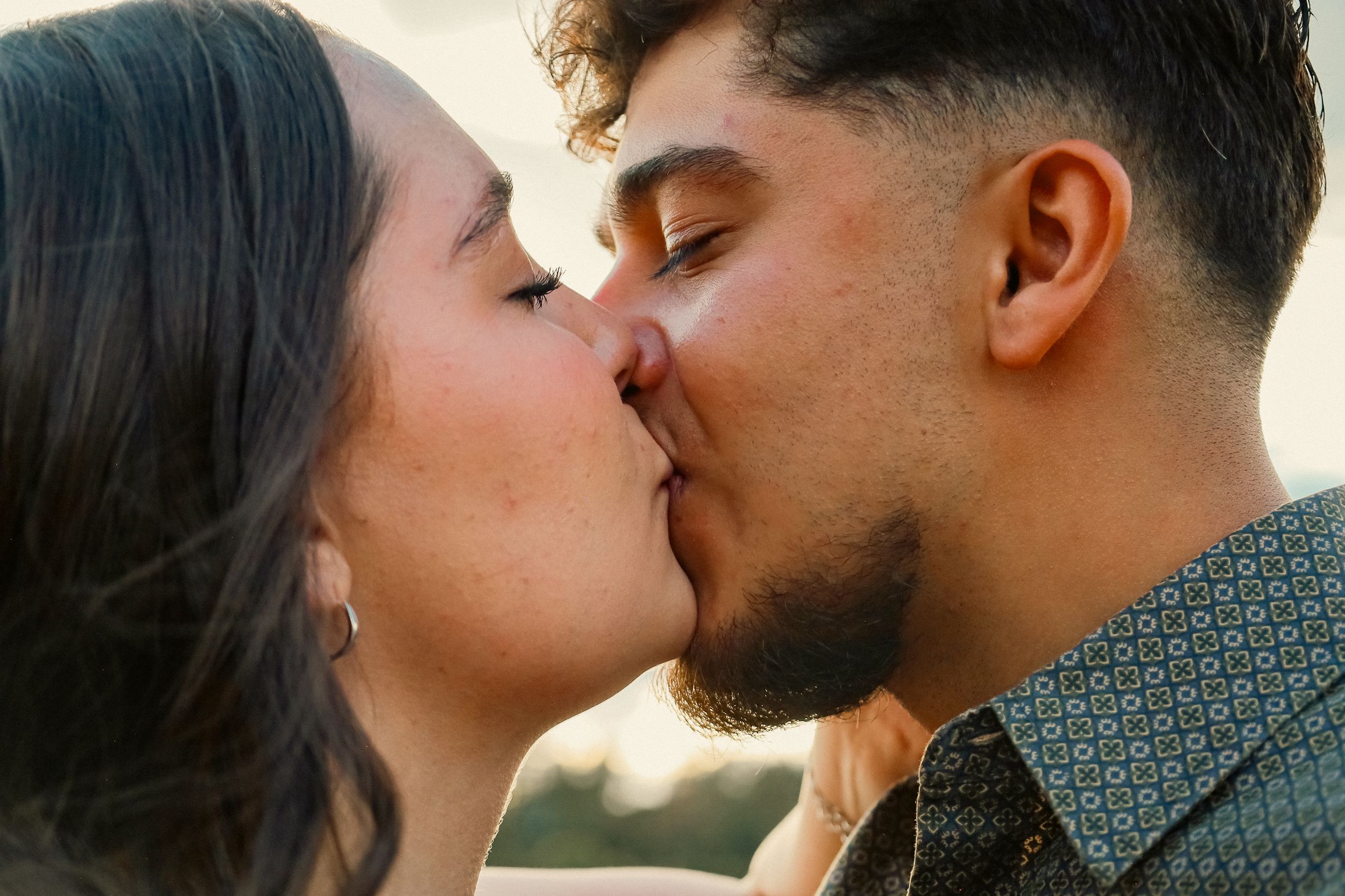 A young couple shares a passionate kiss outdoors in warm golden sunset lighting.
