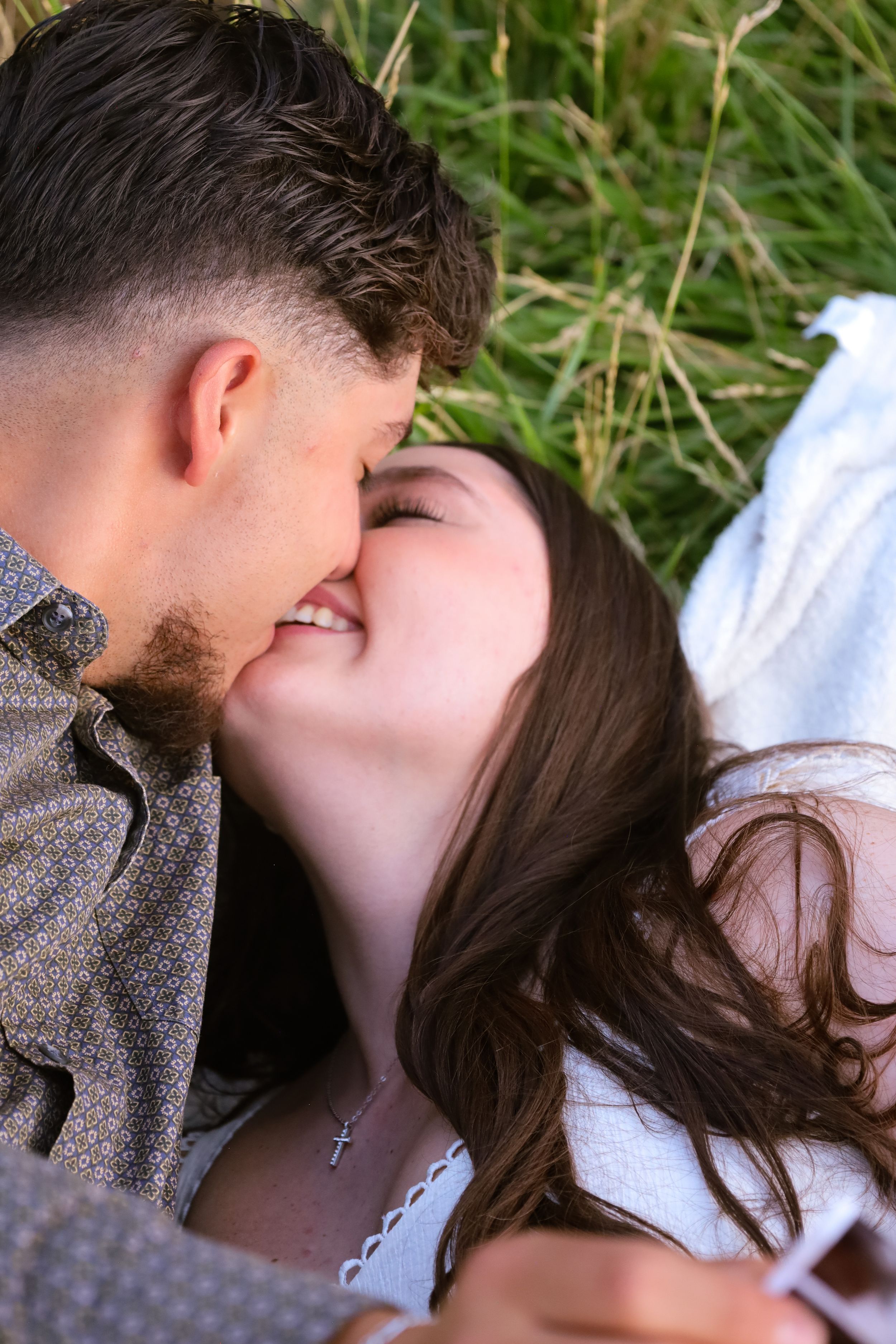 A smiling couple nearly kisses while lying together in a lush green grass field.