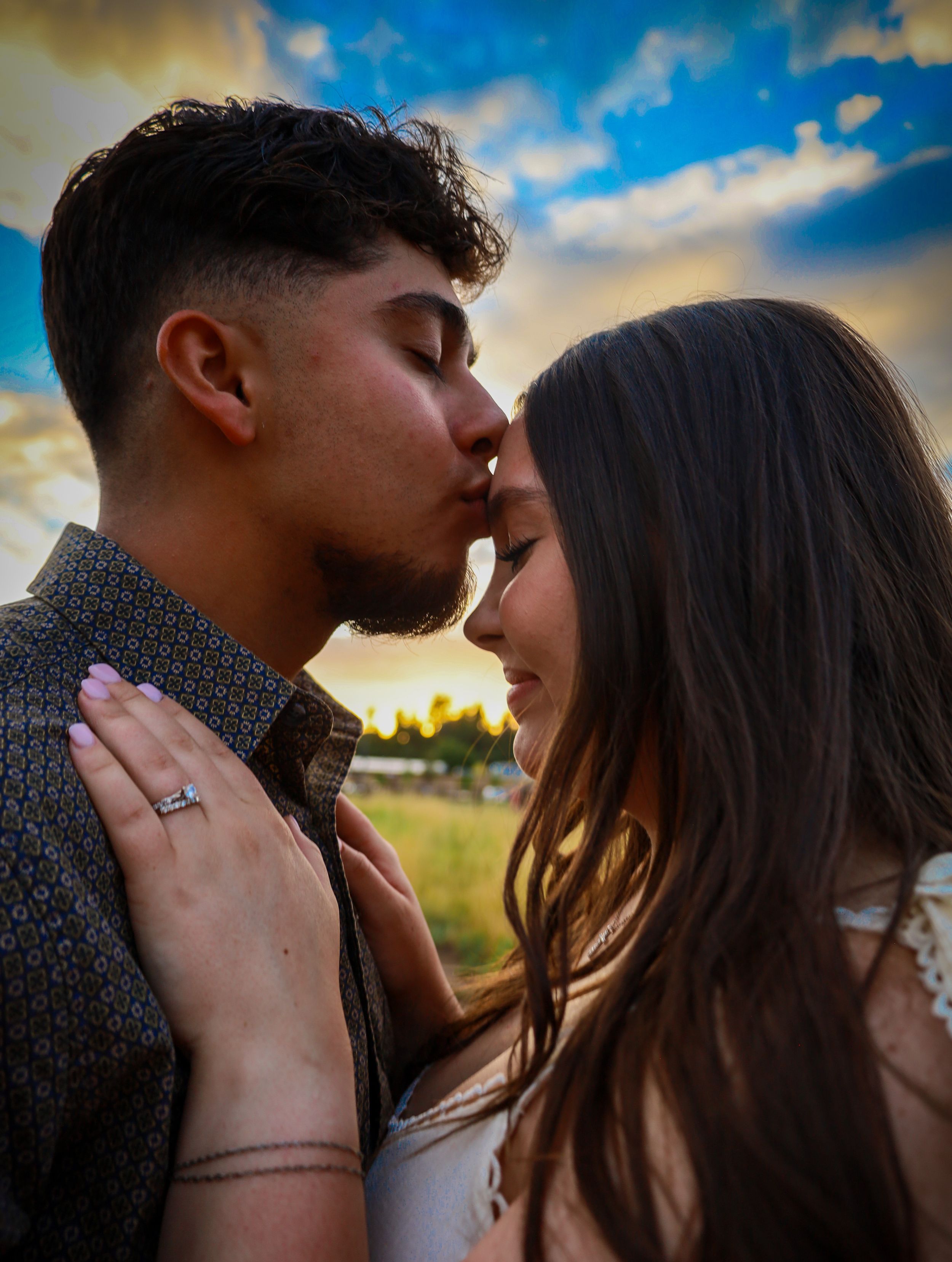 Engaged couple sharing a romantic kiss outdoors at sunset with dramatic cloudy sky.
