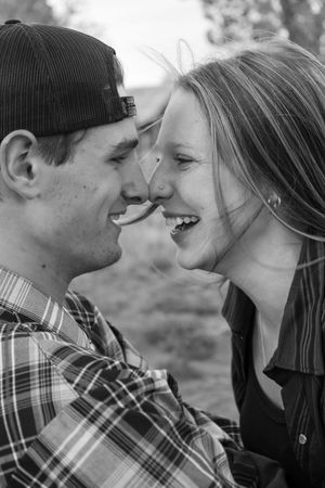 Black and white photo of a laughing young couple touching noses near a rustic barn.