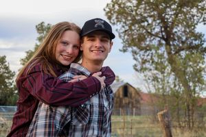 Smiling young couple embracing outdoors with a rustic barn and trees in the background.