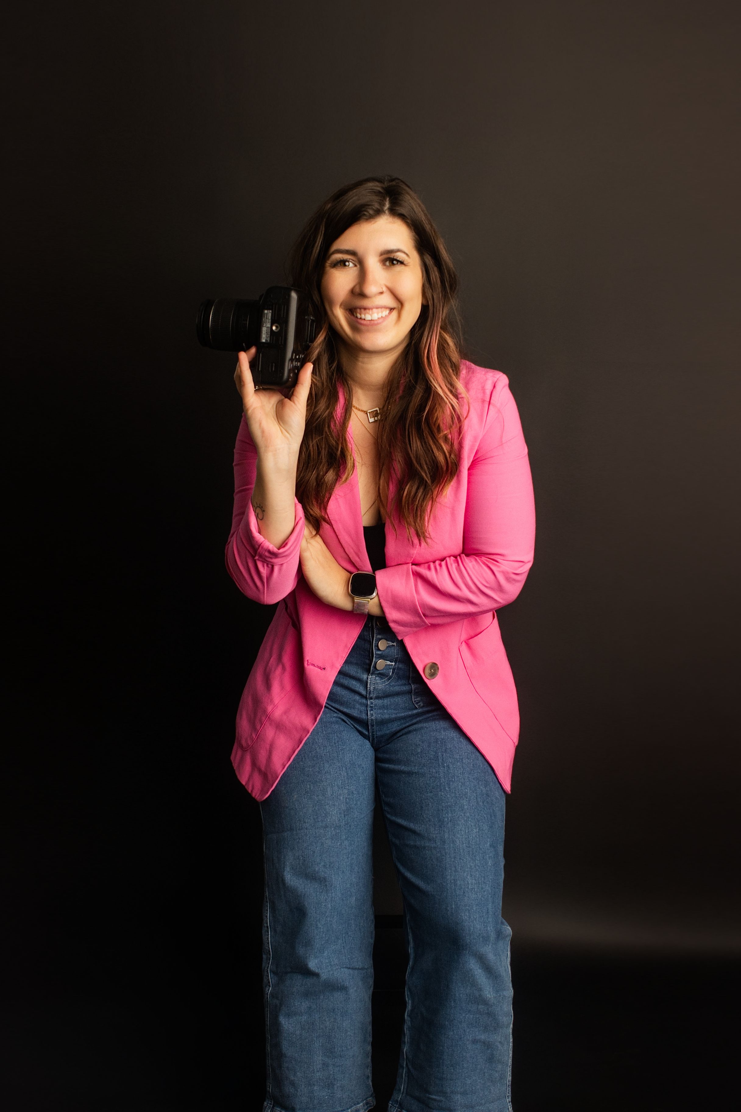 Professional photographer wearing a pink blazer and jeans holds a DSLR camera against dark background.