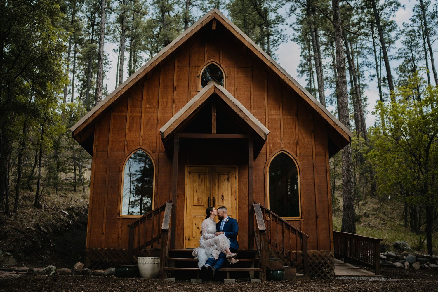 Couple sits on steps of a charming rustic wooden chapel surrounded by pine trees. cinematic wedding photographer new mexico