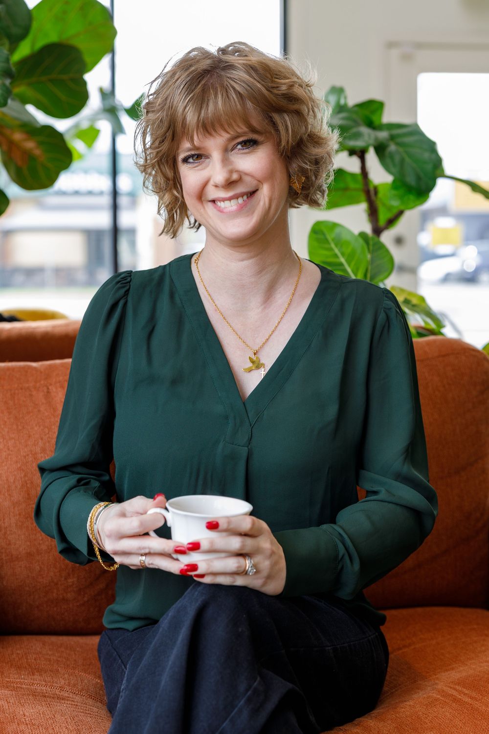 Someone wearing a dark green blouse sits on an orange couch holding a white coffee cup, with houseplants in the background.