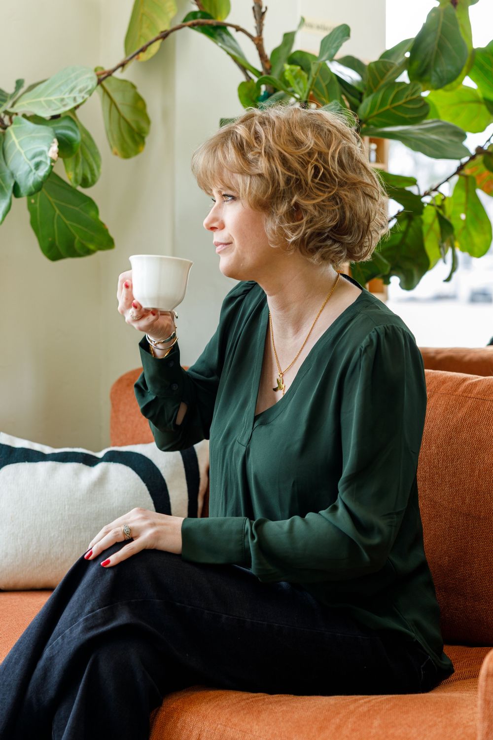 Person in a dark green sweater sits on an orange couch enjoying coffee near a large leafy houseplant.
