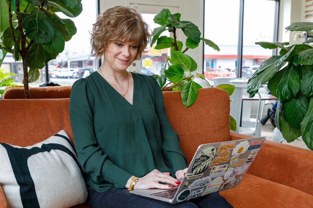A person in dark green attire sits at a desk near large indoor plants in an office setting with orange seating.