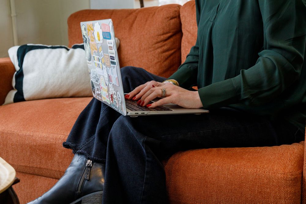 A person wearing green clothing sits on an orange couch while typing on a laptop computer.
