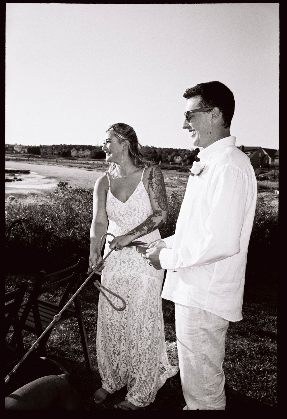 A black and white film photo of a couple laughing together while holding onto their dog who was also their ring bearer in September in Maine