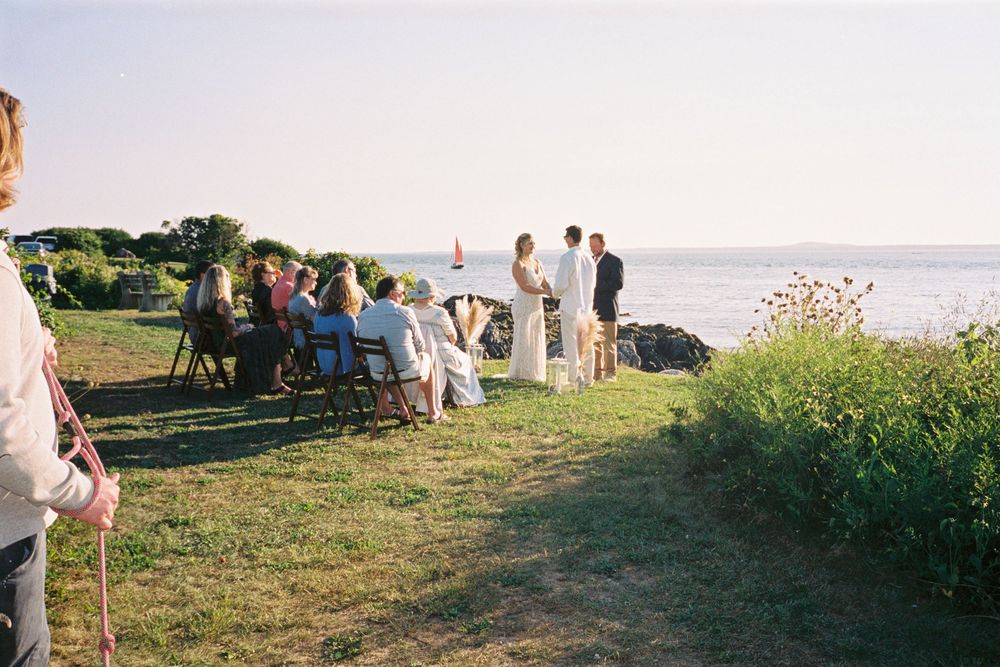 A couple getting married by the sea in a small ceremony with their dogs nearby as a boat sails by in the background on 35mm film in Maine.