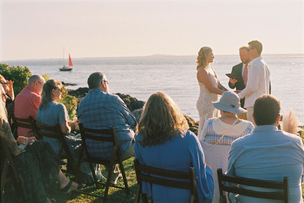 A film photo depicting the guests watching a couple during their small beach ceremony while a boat sails by in the background in the summer.