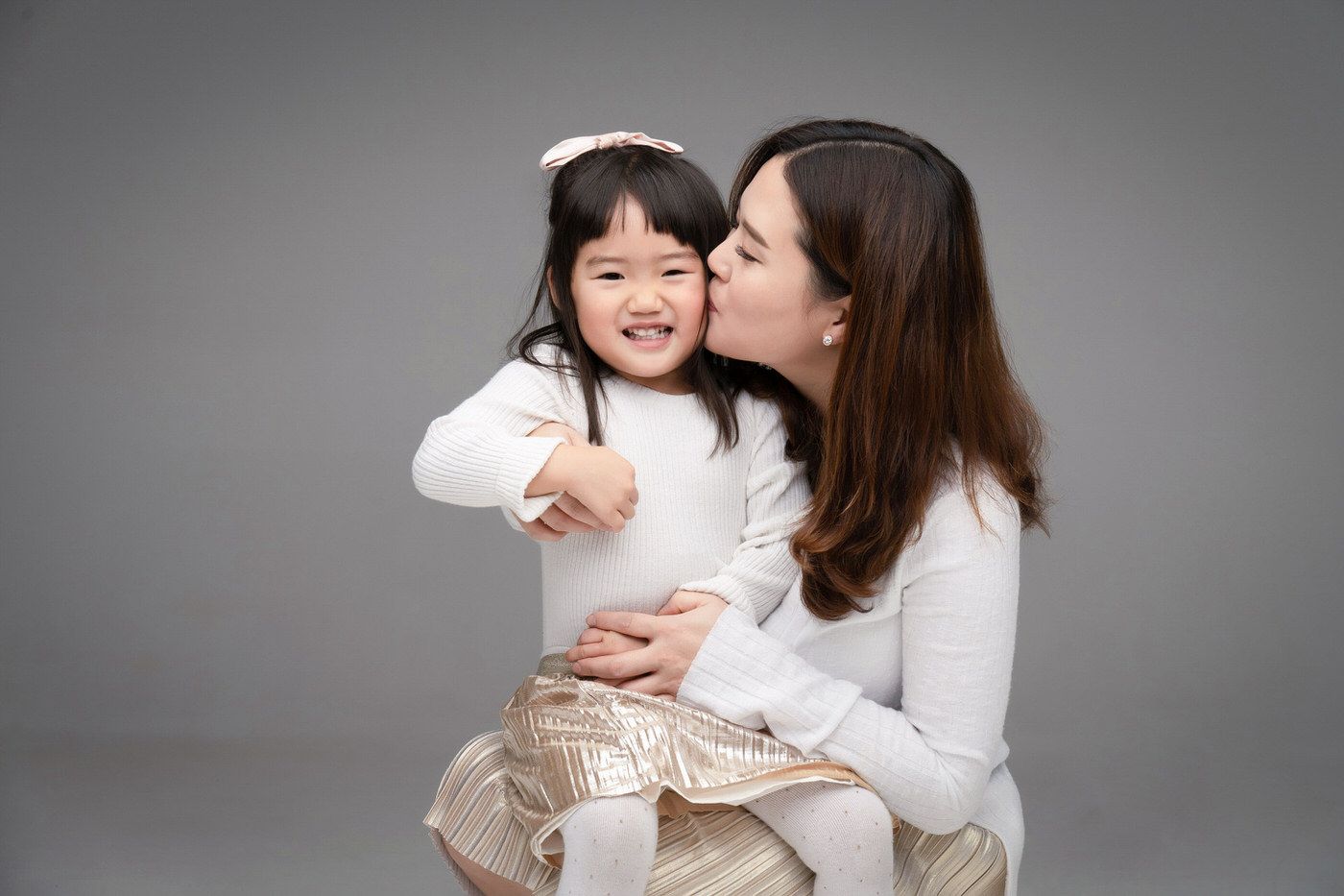 A loving parent in white embraces and kisses a joyful child in matching attire against a gray studio background.